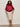 A woman with long curly hair and glasses wears the Sanctuary Clothing sparkle sequin pencil skirt mother of pearl extended sizing, paired with a shiny burgundy zip-up jacket and strappy heels, standing against a plain light wall.