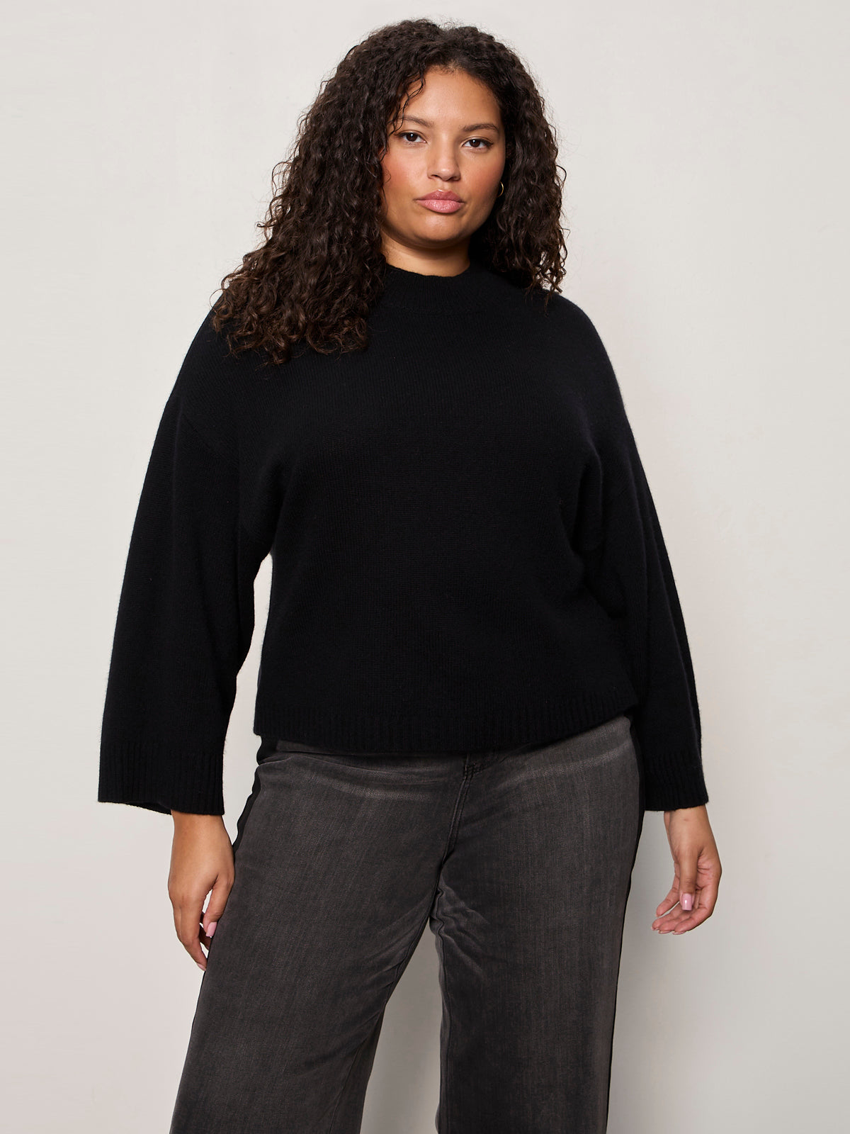 A woman with curly hair models the Sanctuary Clothing forever cashmere popover black extended sizing sweater with gray jeans, confidently standing against a plain light background and looking at the camera.