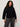 A woman with curly hair models the Sanctuary Clothing forever cashmere popover black extended sizing sweater with gray jeans, confidently standing against a plain light background and looking at the camera.