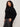 A woman with curly hair confidently models the Sanctuary Clothing forever cashmere popover in black extended sizing, paired with dark grey pants with a black stripe, standing against a light background.