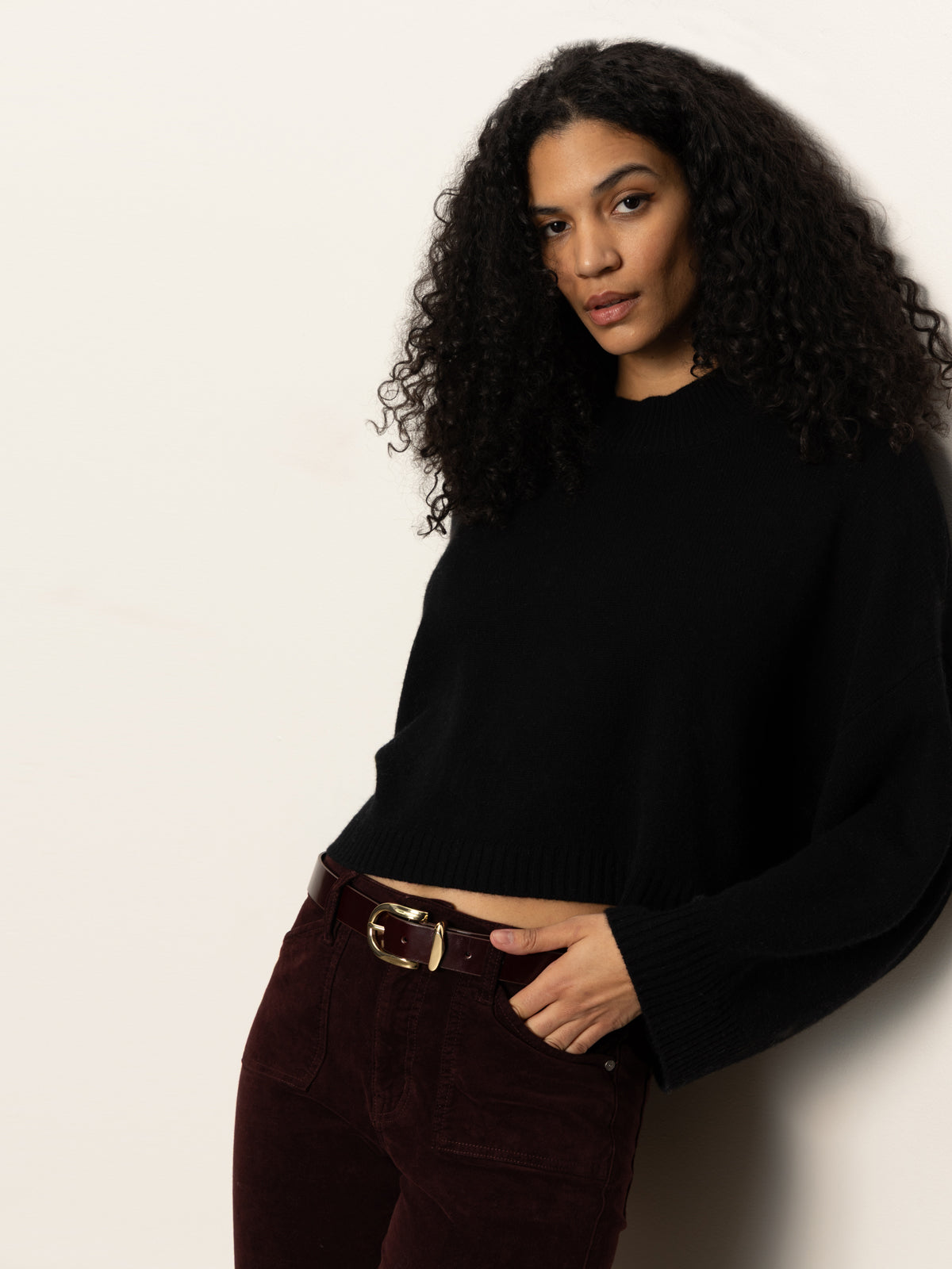 A woman with long curly hair stands confidently against a light background, wearing the Sanctuary Clothing all day belt in dark crimson, paired with a black sweater and her hand in her pocket.