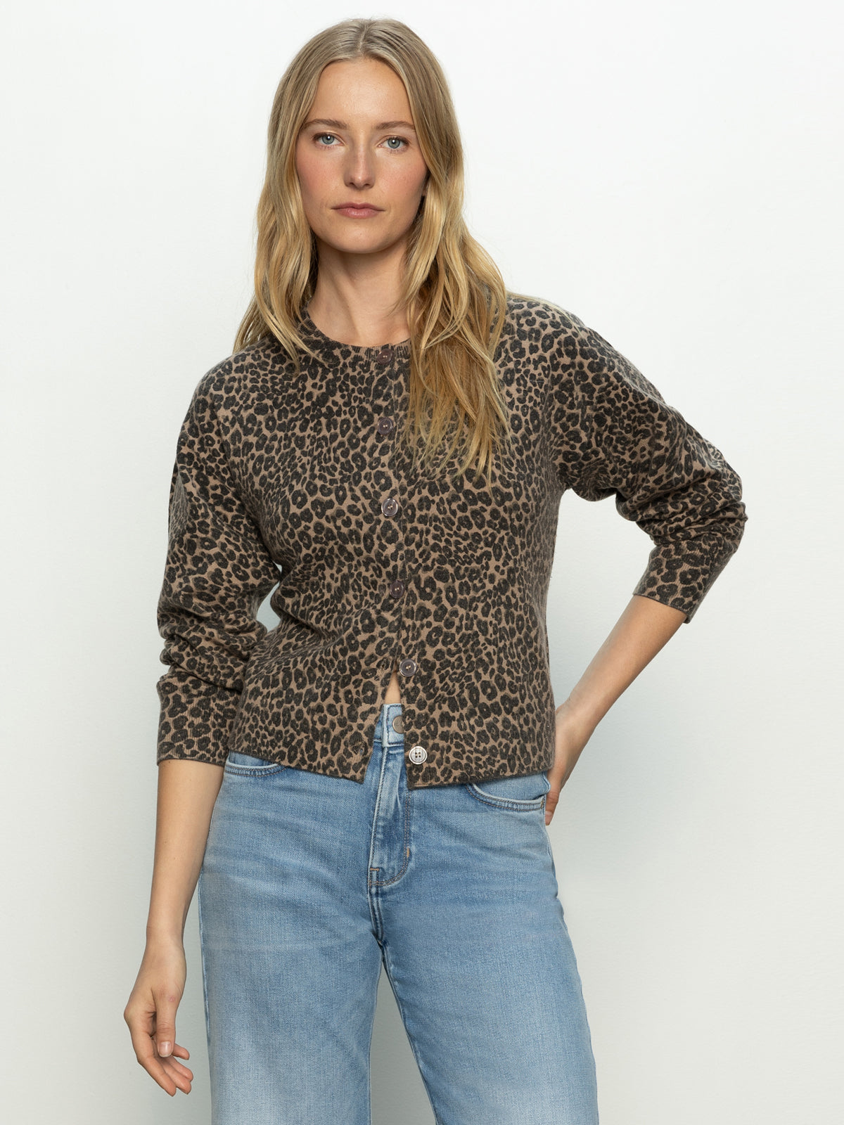 A woman with long blonde hair models the Sanctuary Clothing must have cardi french kitty, a leopard print sweater, styled with light blue jeans against a plain background, one hand in her pocket.