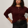 A woman with curly hair models the Sanctuary Clothing boucle cardi in dark crimson (extended sizing), paired with dark high-waisted jeans, standing against a plain light background with one hand on her hip and a neutral expression.