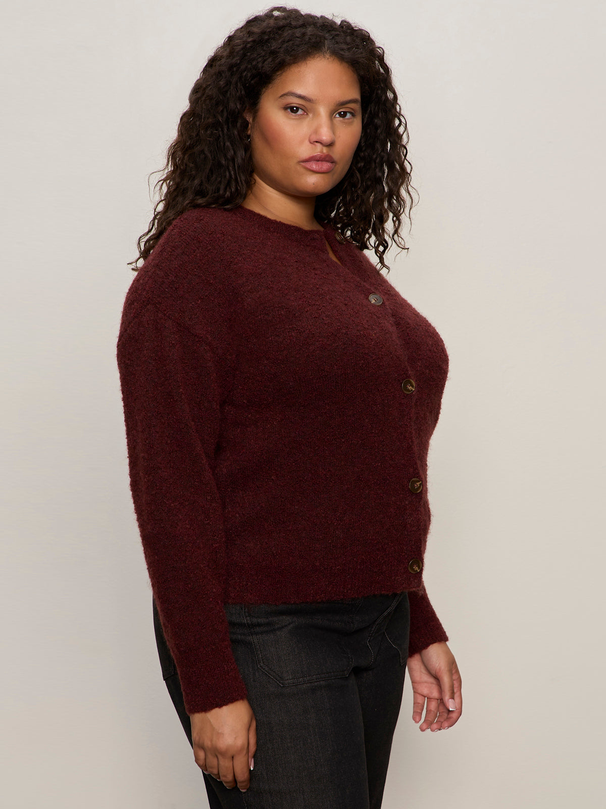 A woman with curly dark hair models the Sanctuary Clothing boucle cardi in dark crimson (extended sizing), paired with dark jeans, standing against a plain light background and looking neutrally at the camera.