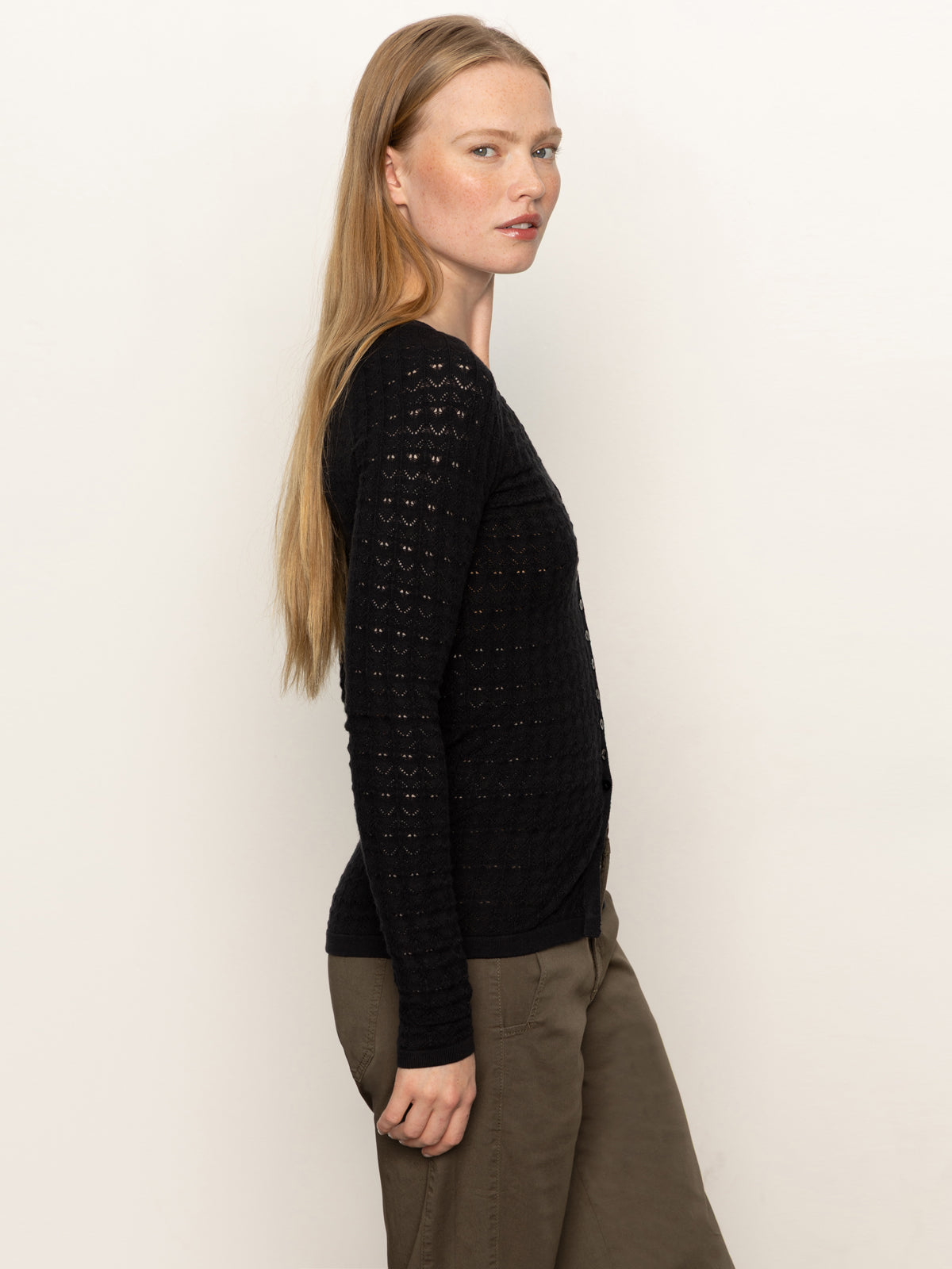 A woman with long, straight blonde hair stands in profile against a plain light background, wearing Sanctuary Clothing's fitted raglan cardi in black and olive green pants, looking slightly over her shoulder.