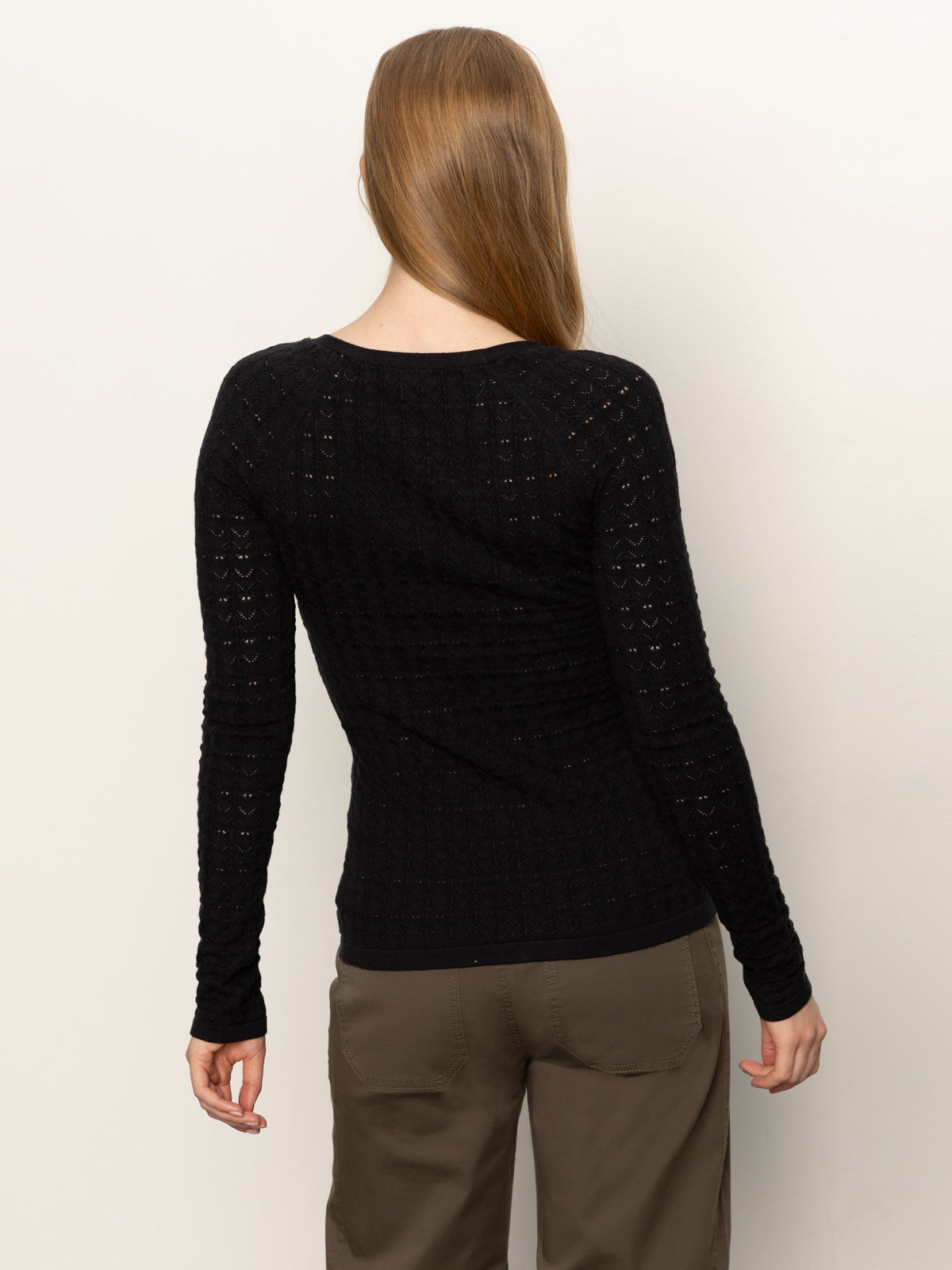 A person with long, straight light brown hair stands with their back to the camera, wearing the Sanctuary Clothing fitted raglan cardi in black and olive-green pants against a plain light background.
