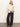 A woman with straight, light hair poses confidently against a plain background, wearing the Sanctuary Clothing fitted raglan cardi in light oat, paired with wide-leg black pants and dark ankle boots.