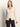 A woman with straight, light brown hair wears the Sanctuary Clothing fitted raglan cardi in light oat—a cream-colored, long-sleeve V-neck cardigan—paired with dark pants. She stands against a plain, light background, looking slightly to the side.