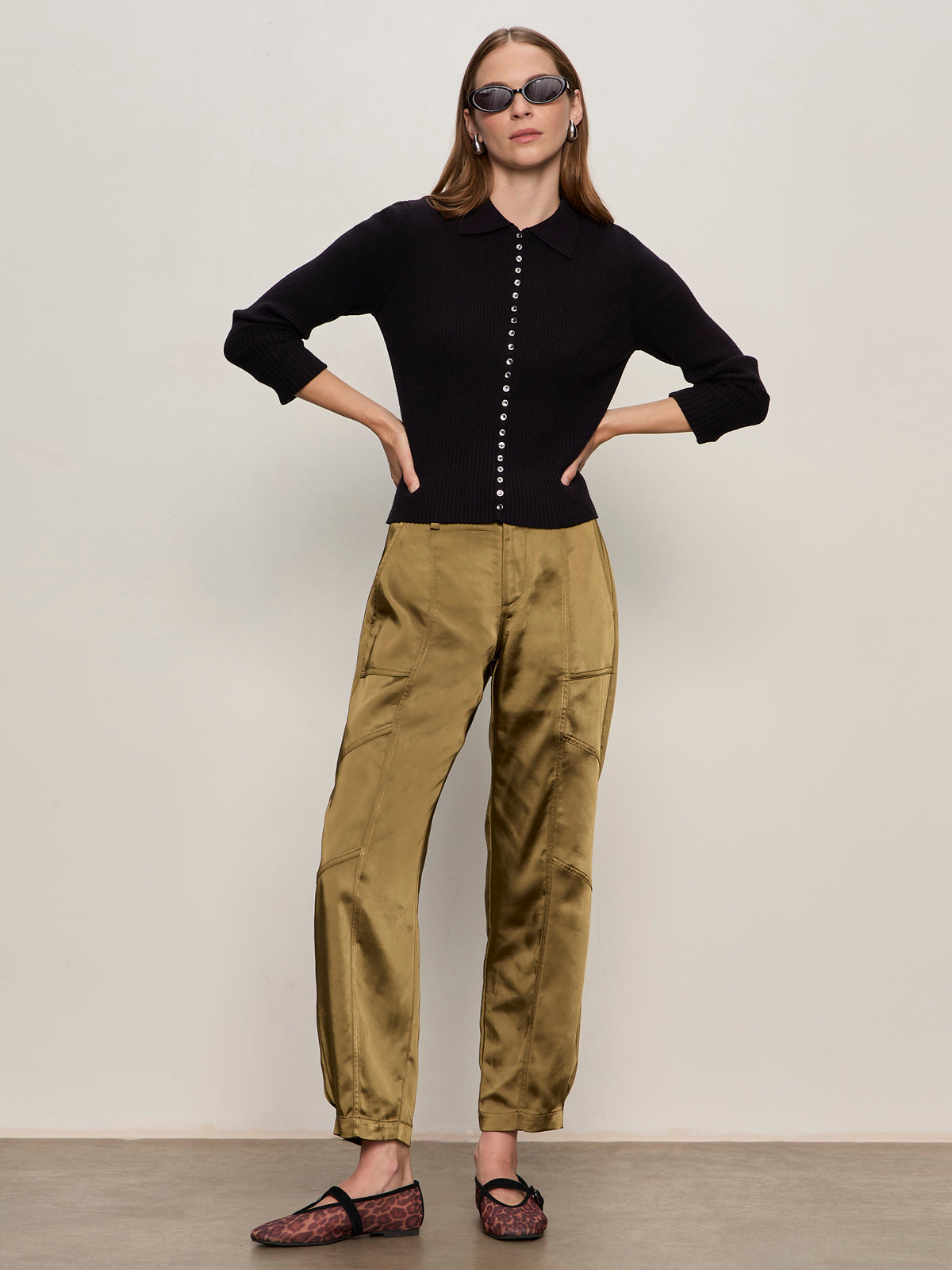 A woman stands against a light wall with her hands on her hips, wearing black sunglasses, Sanctuary Clothing's button down variegated cardi in black, khaki satin pants, and animal-print flats.