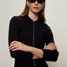 A woman with straight brown hair wears oval black sunglasses, silver hoop earrings, and the Sanctuary Clothing button down variegated cardi in black. She stands with arms crossed against a plain light background.
