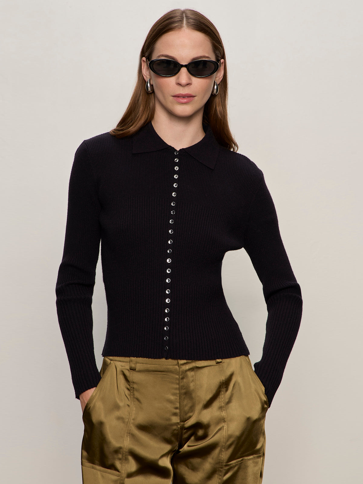 A woman with long brown hair wears black oval sunglasses, shiny gold pants, and the Sanctuary Clothing button down variegated cardi black, standing against a plain light background with her hands in her pockets.
