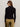 A person with long brown hair, wearing black sunglasses and the Sanctuary Clothing button down variegated cardi in black, stands facing a plain light-colored wall, shown from the back.