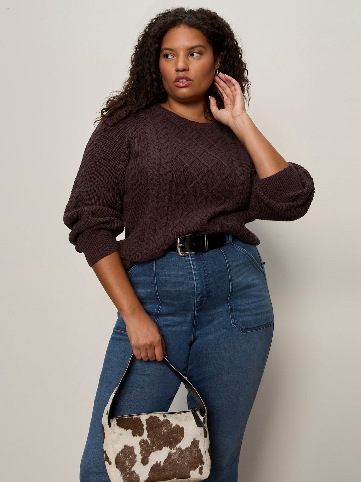 A woman with curly hair wears a brown cable-knit sweater, high-waisted blue jeans, and the Sanctuary Clothing rowan belt in black. She holds a small cow-print handbag and stands against a plain light background.