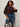 A woman with curly hair wears a brown cable-knit sweater, high-waisted blue jeans, and the Sanctuary Clothing rowan belt in black. She holds a small cow-print handbag and stands against a plain light background.