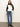 A woman with long wavy hair and a neutral expression stands against a plain wall, wearing Sanctuary Clothing’s downtown mock neck sweater in black chalk, paired with blue jeans and black ankle boots. One hand is by her side, the other in her pocket.
