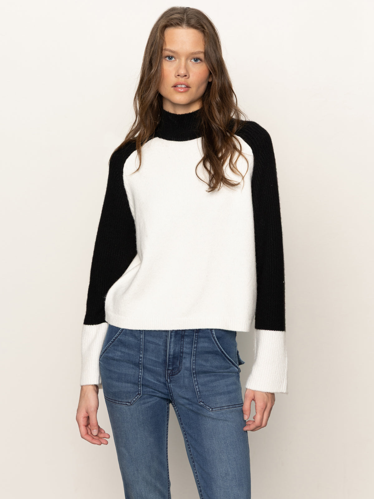 A woman with long brown hair wears the Sanctuary Clothing downtown mock neck sweater in black chalk and blue jeans, standing against a plain light background and looking directly at the camera.