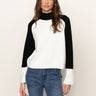 A woman with long brown hair wears the Sanctuary Clothing downtown mock neck sweater in black chalk and blue jeans, standing against a plain light background and looking directly at the camera.