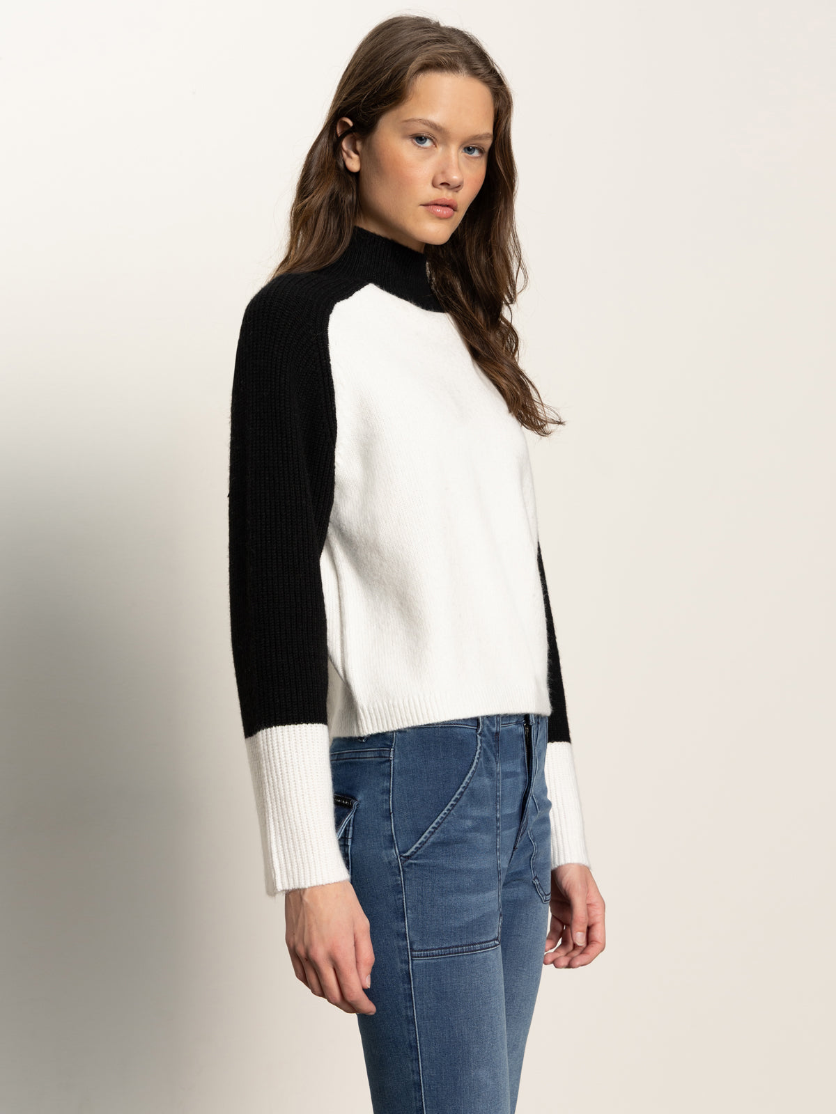 A woman with long brown hair models the Sanctuary Clothing downtown mock neck sweater in black chalk with blue jeans, standing against a plain light background and gazing slightly to the side.