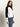 A woman with long brown hair models the Sanctuary Clothing downtown mock neck sweater in black chalk with blue jeans, standing against a plain light background and gazing slightly to the side.