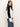 A woman with long brown hair wears the Sanctuary Clothing downtown mock neck sweater in black chalk, paired with blue jeans, standing sideways against a plain light-colored background.