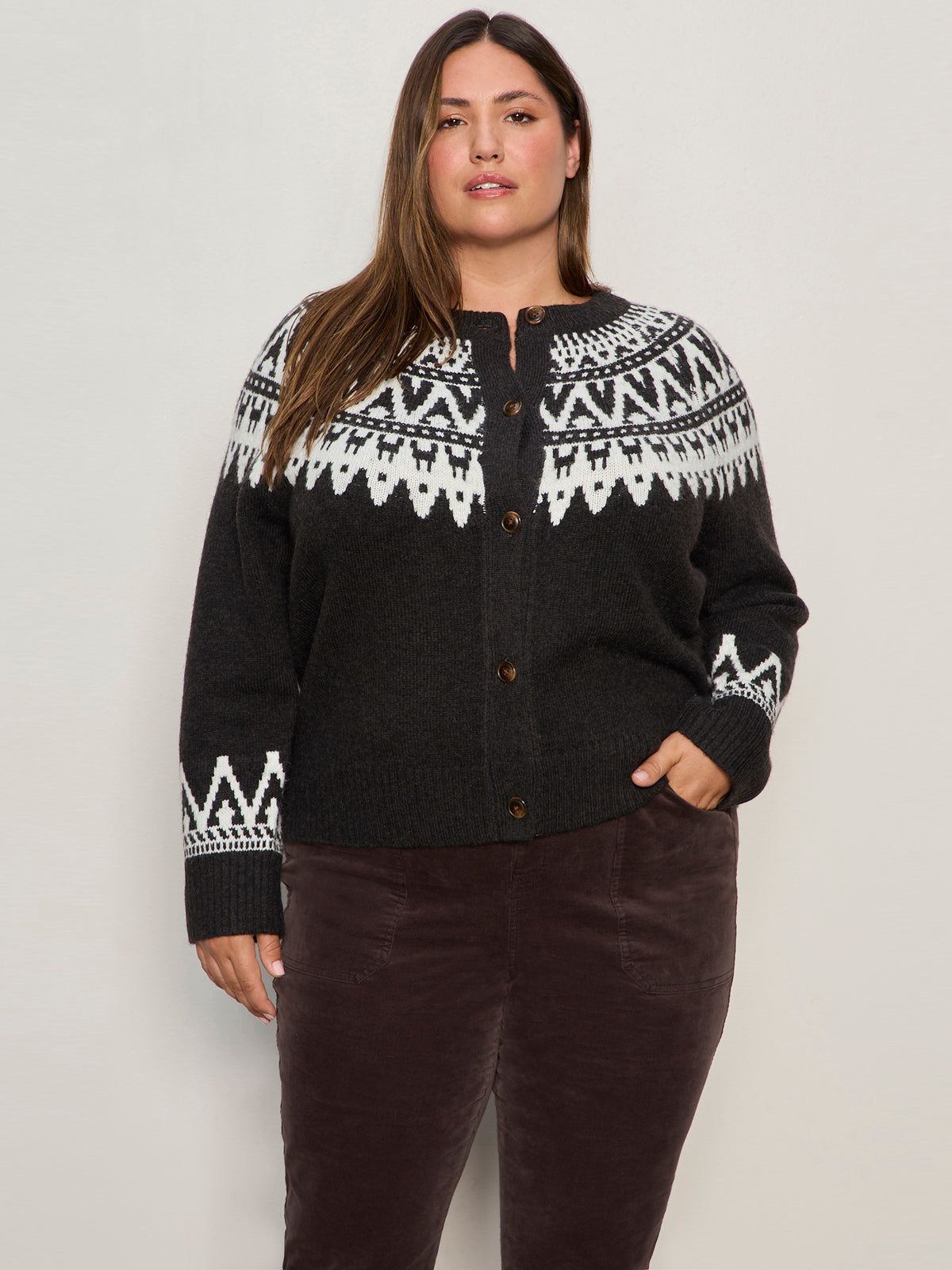 A woman with long brown hair models the Sanctuary Clothing fairisle crewneck cardi in ink multi (extended sizing), featuring a black base with white geometric patterns. She stands against a light background, one hand in her pocket.