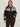 A woman with long brown hair models the Sanctuary Clothing fairisle crewneck cardi in ink multi (extended sizing), featuring a black base with white geometric patterns. She stands against a light background, one hand in her pocket.