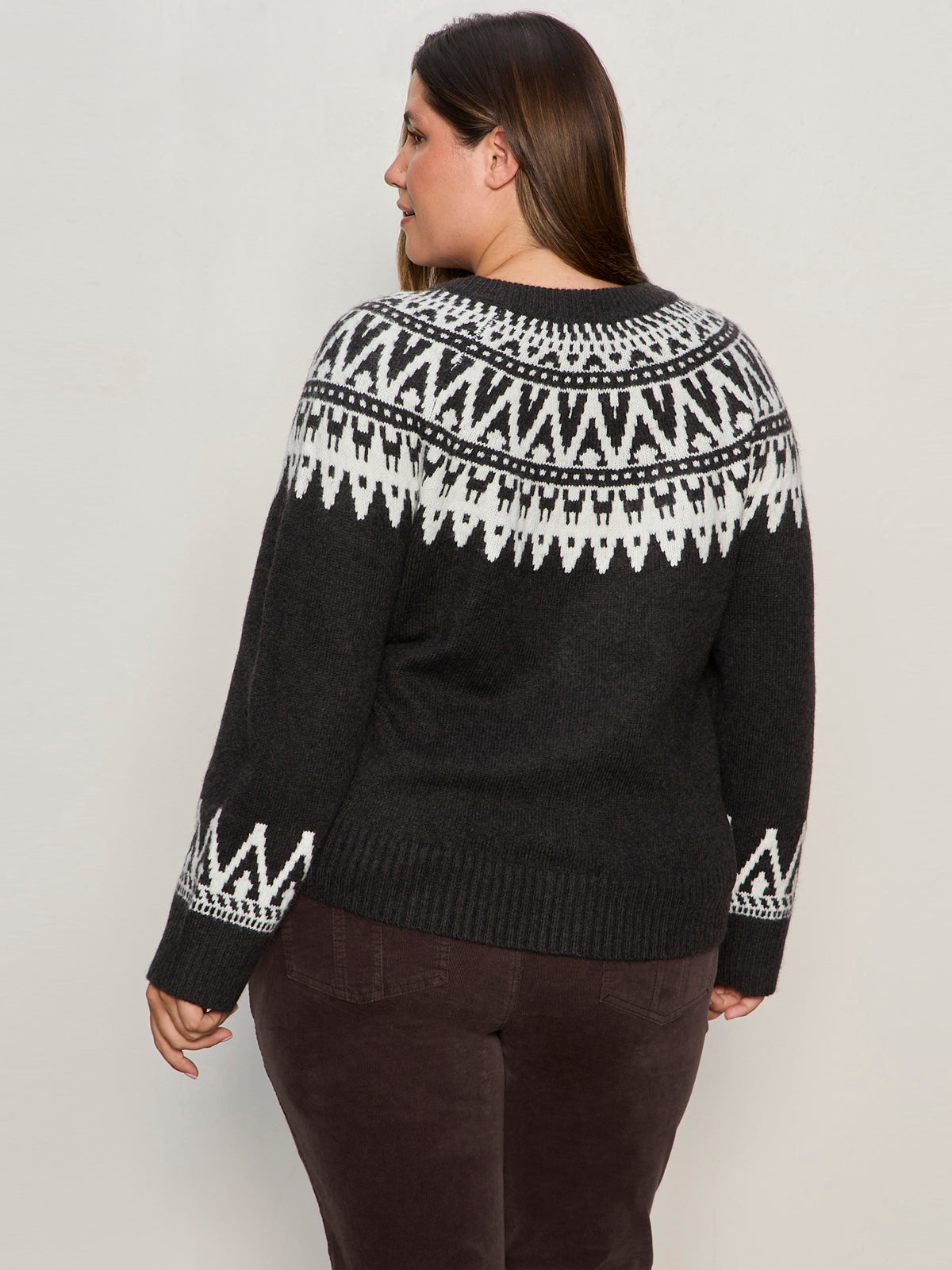 A woman with long brown hair, dressed in Sanctuary Clothing’s fairisle crewneck cardi ink multi extended sizing and dark brown pants, stands facing away and looks over her left shoulder.