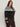 A woman with long brown hair wears the Sanctuary Clothing fairisle crewneck cardi ink multi extended sizing sweater, paired with brown corduroy pants, standing against a plain light background.