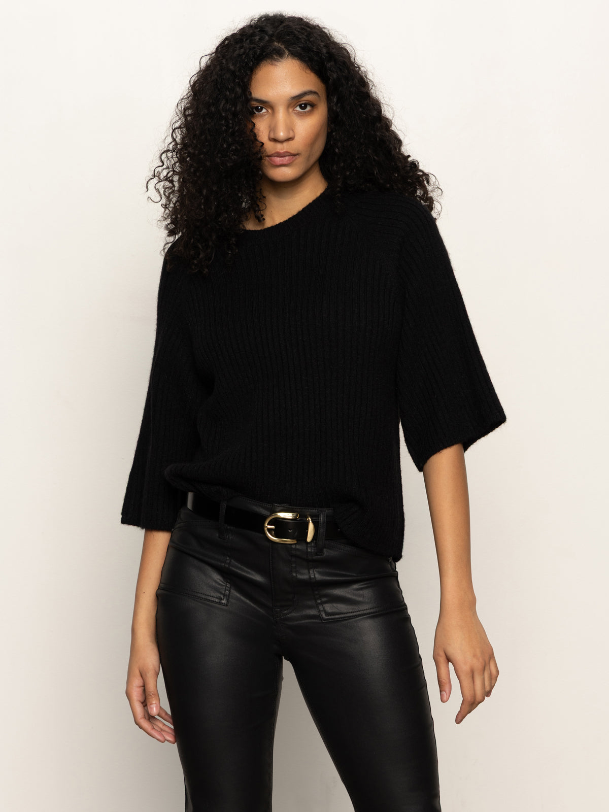 A woman with curly hair wears the Sanctuary Clothing all day belt in black, paired with a black knit sweater and black leather pants, standing against a plain light background and looking directly at the camera.