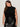 A woman with long brown hair wears the Sanctuary Clothing city sweater shell in black extended sizing and a high-waisted black leather skirt, standing against a plain light background, looking down with a gentle expression.