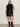 A woman wears the Sanctuary Clothing city sweater shell in black (extended sizing) with a leather skirt and boots, standing confidently against a plain light wall.
