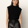 A woman with wavy brown hair wears the Sanctuary Clothing city sweater shell in black, paired with high-waisted black leather pants, standing against a light background and looking at the camera with a neutral expression.