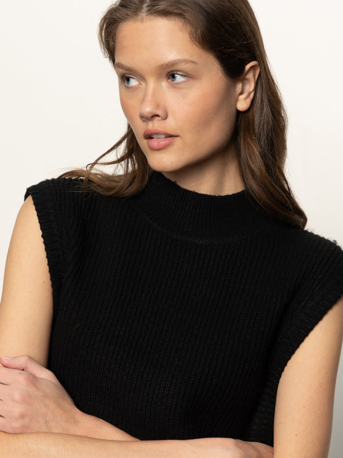 A woman with long brown hair crosses her arms and looks left, wearing the Sanctuary Clothing city sweater shell in black. The sleeveless, high-neck knit top stands out against a plain, light background.