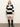 A woman stands against a plain wall wearing the Sanctuary Clothing polo striped sweater dress in heather pearl with black, featuring a white collared neckline and knee-high black boots. She faces forward, hands by her sides.