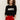 A woman with curly hair wears the Sanctuary Clothing "life of the party" cashmere sweater in black with chalk lettering, paired with bright red pants, standing against a plain light background with her hands in her pockets.