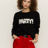 A woman with curly hair wears the Sanctuary Clothing "life of the party" cashmere sweater in black with chalk lettering, paired with bright red pants, standing against a plain light background with her hands in her pockets.