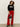 A person with curly hair wears the Sanctuary Clothing "life of the party" black cashmere sweater with œPARTY!� in chalk lettering, paired with bright red pants and black heeled boots, holding a small black purse against a plain background.