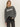 A woman with long blonde hair wears the Sanctuary Clothing "life of the party" cashmere sweater in heather charcoal with chalk lettering, styled with dark jeans and a black belt, standing against a plain light background.