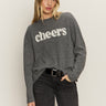 A woman with long blonde hair wears the Sanctuary Clothing "life of the party" cashmere sweater in heather charcoal with chalk lettering, styled with dark jeans and a black belt, standing against a plain light background.