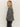 A person with long blonde hair stands in profile, wearing the Sanctuary Clothing 'life of the party' cashmere sweater in heather charcoal with chalk, paired with dark jeans and hands in pockets, against a plain light background.