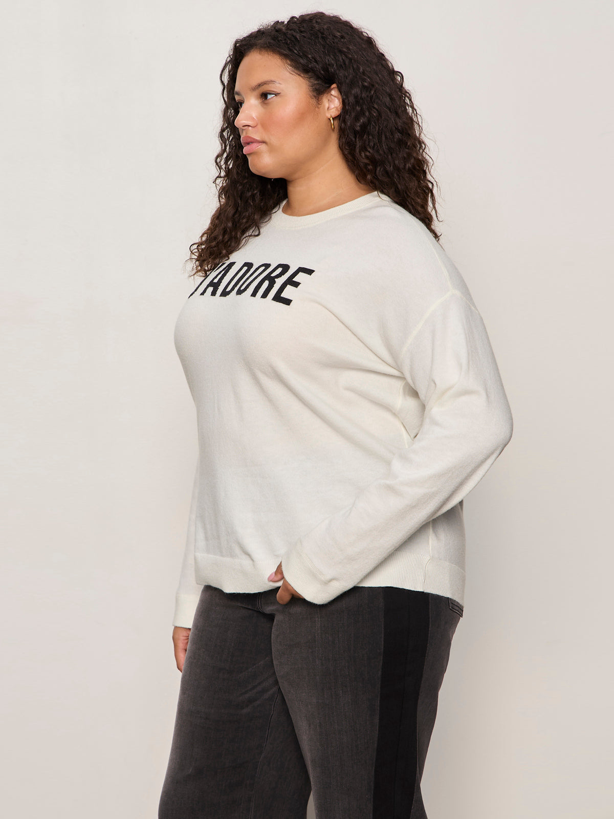 A woman with curly hair stands sideways, wearing a white long-sleeve sweater with the word "AMORE" in black letters and dark pants, against a plain light background.