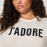 A woman with curly brown hair wears a cream-colored sweater with the word "J'ADORE" in bold black letters on the front. She has a neutral expression and is facing the camera.