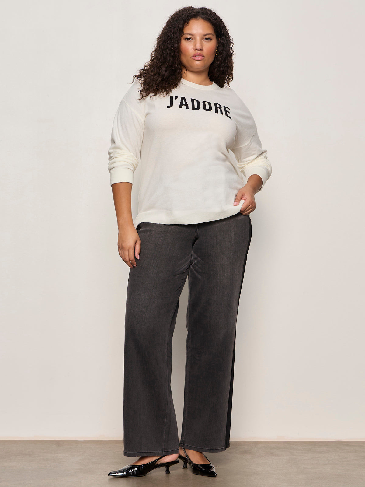 A woman with curly hair wears a cream sweater with "J'ADORE" written on it, dark wide-leg jeans, and black shoes, standing against a plain light background.