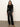 A woman with loose, wavy hair and a neutral expression wears the Sanctuary Clothing long sleeve shoulder twist top in black, paired with high-waisted black leather pants and tan pointed-toe boots against a light background.