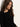 A young woman with long wavy brown hair, wearing the Sanctuary Clothing long sleeve shoulder twist top in black, stands against a light-colored wall and looks down calmly.