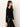 A woman with long brown hair wears the Sanctuary Clothing long sleeve shoulder twist top in black, styled with black leather pants and a studded belt, standing in profile against an off-white background.