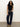 A woman with curly hair wears the Sanctuary Clothing asymm shoulder twist top in black, paired with dark blue wide-leg jeans, a leopard print belt, and tan ankle boots against a plain light-colored background.