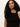 A woman with long, curly dark hair wears the Sanctuary Clothing asymm shoulder twist top in black, standing against a plain light background and looking confidently at the camera.