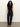 A woman with long curly hair wears a black sleeveless top, high-waisted dark blue jeans with white stitching, and tan heeled boots, standing against a plain light-colored background.