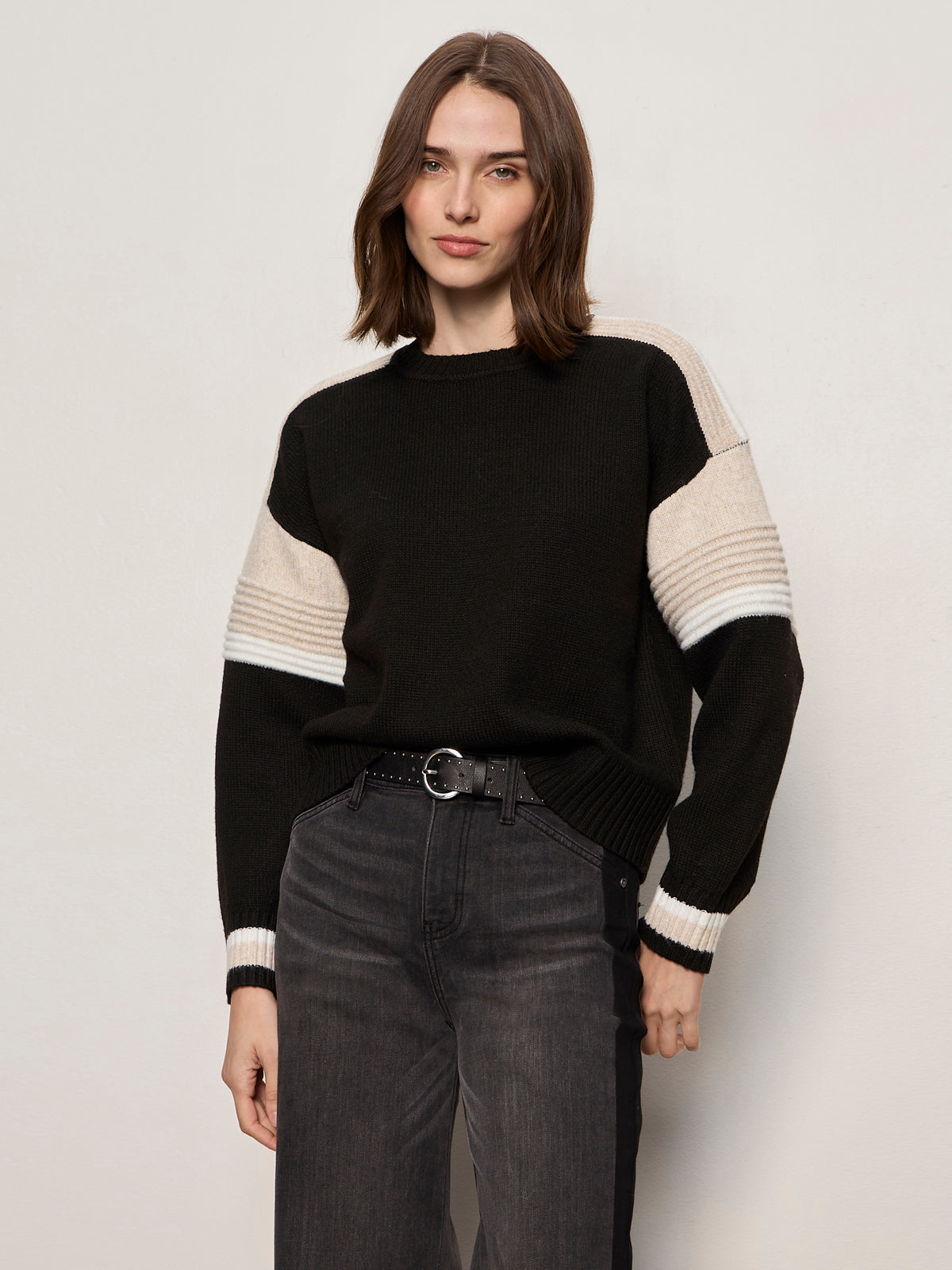 A person with shoulder-length brown hair wears the Sanctuary Clothing sporty stripe crewneck sweater in black multi and dark gray jeans, standing against a plain light-colored wall.